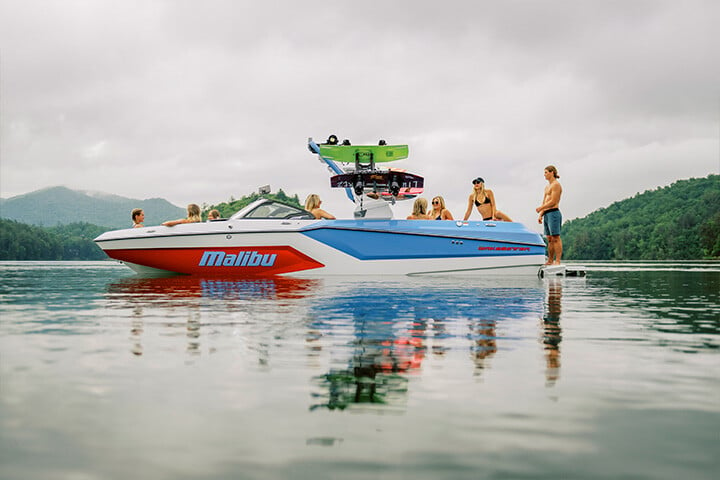 A red, white, and blue All-New 2025 Malibu Wakesetter 24 MXZ sitting in the water with friends relaxing throughout the lounge