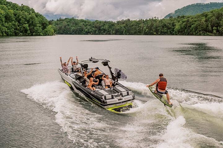 A person jumping and doing a trick on a wakesurf board behind a black, gray, and yellow 2025 Malibu Boats Wakesetter 23 MXZ A person jumping and doing a trick on a wakesurf board behind a black, gray, and yellow 2025 Malibu Boats Wakesetter 23 MXZ